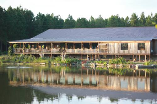 A wooden building with a porch reflects in a calm lake, surrounded by trees and greenery.