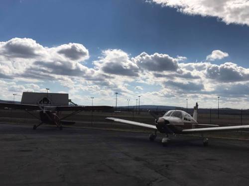 Two small airplanes parked on a tarmac under a cloudy sky, with a distant building and mountains in the background.