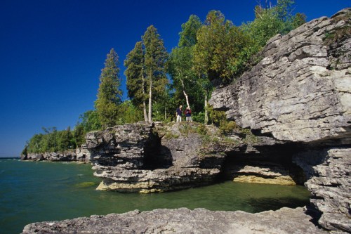 Two people stand on a rocky shoreline with trees, overlooking a calm blue lake under a clear sky.