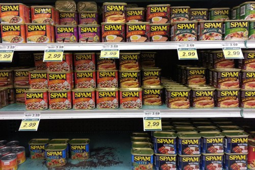 A grocery store shelf filled with various flavors of Spam cans, each labeled with prices.