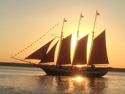 A tall ship with red sails sails on calm waters at sunset, with the sun glowing behind it and flags fluttering.