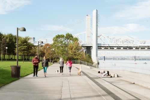People walking dogs along a riverside path with a bridge in the background and trees lining the walkway.
