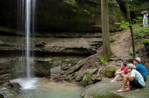 A group of people sits by a waterfall in a lush forest, enjoying the serene natural surroundings.