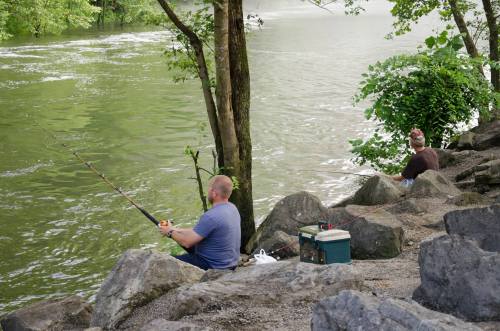 Two people fishing by a river, sitting on rocks with a cooler nearby, surrounded by trees and greenery.
