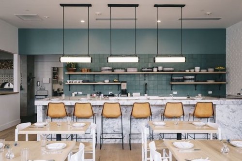 Modern restaurant interior featuring a marble bar, leather stools, and a stylish kitchen backdrop with shelves and table settings.