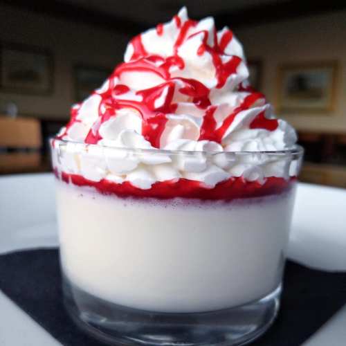 A glass dessert topped with whipped cream and red syrup, layered with a creamy base and a fruit sauce.