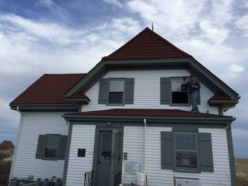 A person stands on the roof of a white house with a red roof, working on a window under a cloudy sky.