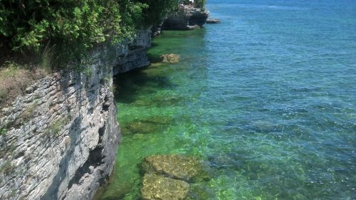 Clear turquoise water gently laps against rocky cliffs, with lush greenery framing the serene shoreline.