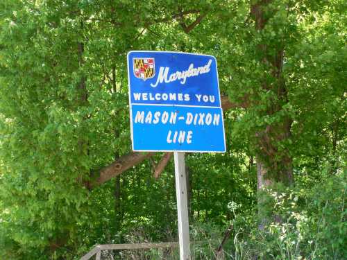 A blue welcome sign for Maryland marking the Mason-Dixon Line, surrounded by green trees.