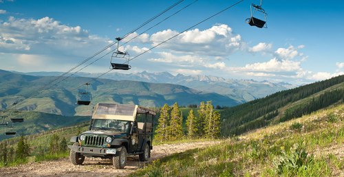 A rugged vehicle drives along a mountain path under a ski lift, surrounded by lush greenery and distant peaks.