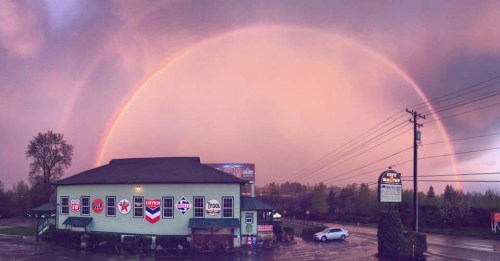 A colorful rainbow arcs over a gas station with various brand signs, set against a cloudy sky.