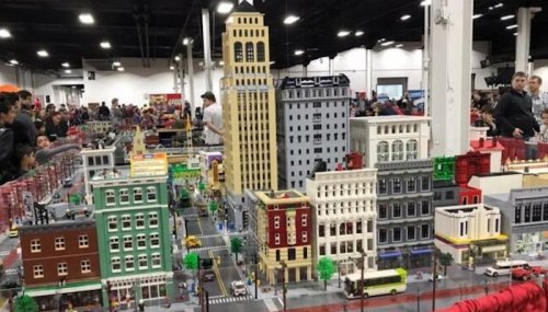 A detailed LEGO cityscape with tall buildings, streets, and crowds of people in a large exhibition space.