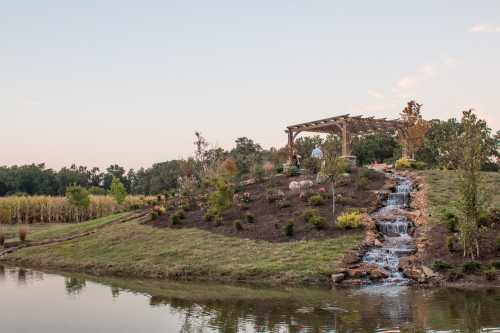 A serene landscape featuring a pond, a waterfall, and a garden with a pergola on a hill at sunset.