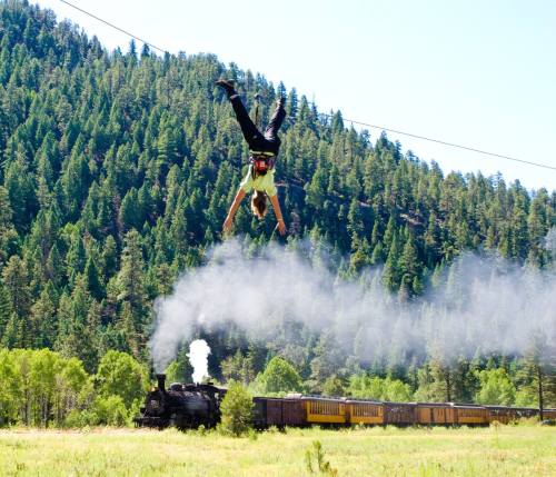 A person hangs upside down from a zip line above a train traveling through a forested landscape.