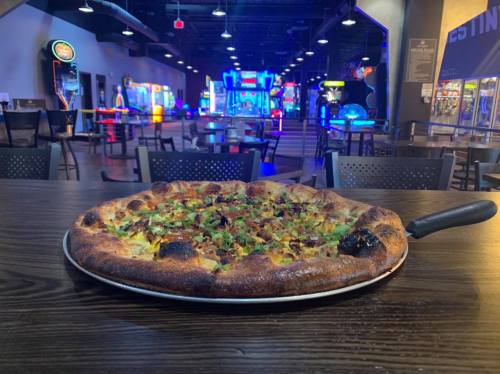 A freshly baked pizza with toppings sits on a table, with an arcade in the background illuminated by colorful lights.