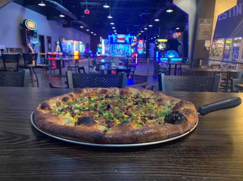 A freshly baked pizza with toppings sits on a table, with an arcade in the background illuminated by colorful lights.