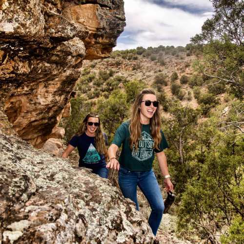 Two women climbing a rocky trail, smiling and wearing sunglasses, surrounded by greenery and rugged terrain.