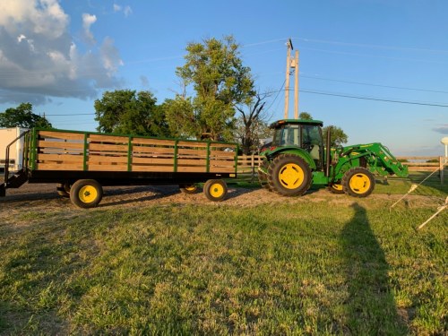 A green tractor with a wooden trailer parked on a grassy field under a blue sky with scattered clouds.
