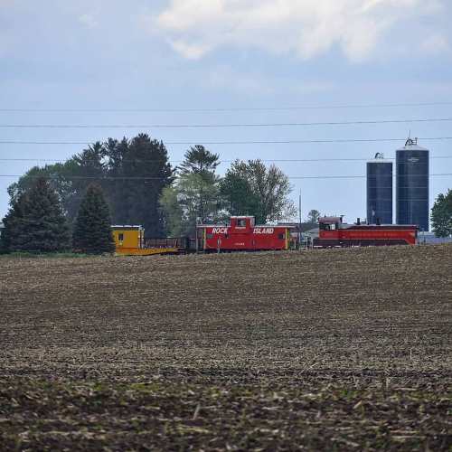 A rural landscape featuring fields, a train, and silos under a cloudy sky.