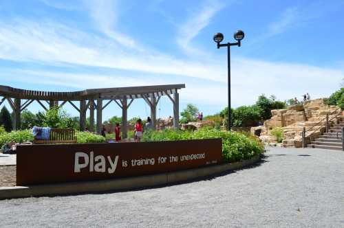 A park scene featuring a sign that reads "Play is training for the unexpected," with greenery and a rocky area in the background.