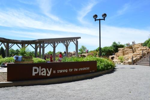 A park scene featuring a sign that reads "Play is training for the unexpected," with greenery and a rocky area in the background.