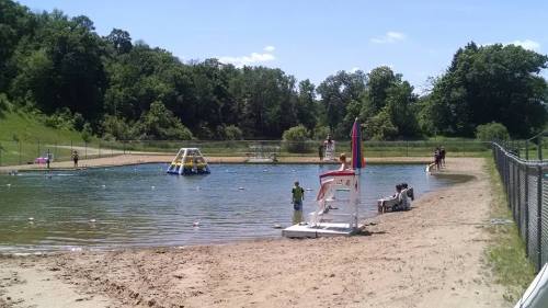 A sandy beach area by a swimming pond, with people swimming, a lifeguard tower, and green trees in the background.