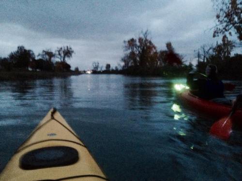 Two kayaks on a calm, dark waterway under a cloudy sky, with trees and distant lights in the background.