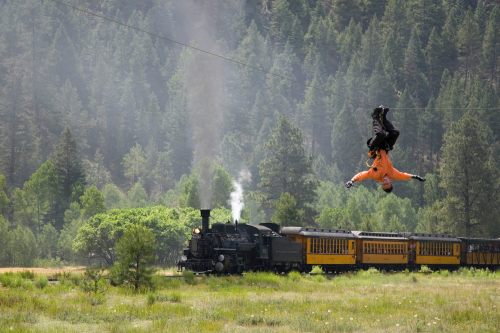 A person zip-lining above a train in a lush, green landscape with trees and mountains in the background.