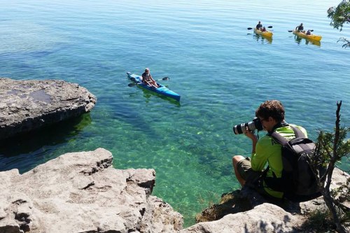 A person sits on a rock, photographing two kayakers in clear blue water on a sunny day.