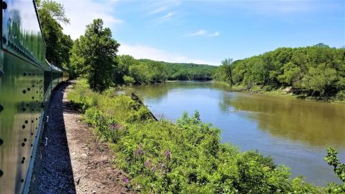 A scenic view of a river bordered by lush greenery, with a train track running alongside under a clear blue sky.