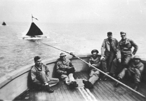 A group of six men in military attire sitting on a boat, with a sailboat visible in the background on a cloudy day.