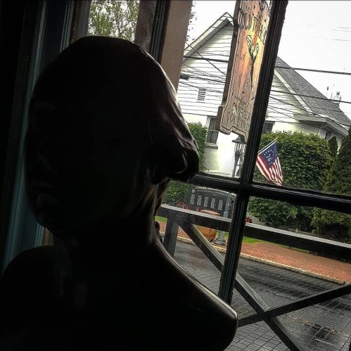 A dark silhouette of a bust in front of a window showing an American flag and a house in the background.