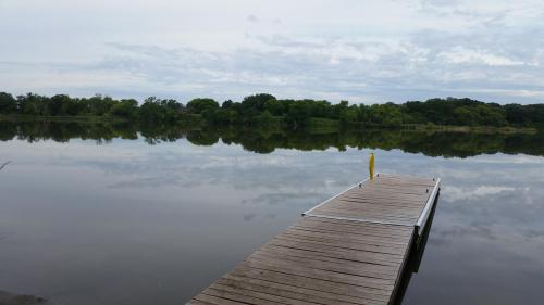 A calm lake with a wooden dock extending into the water, surrounded by lush greenery and a cloudy sky.