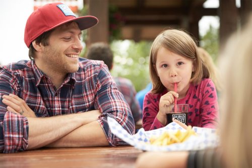A man and a young girl share a meal outdoors, smiling as she drinks from a cup with a straw. French fries are on the table.