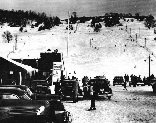 A snowy ski slope with skiers, vintage cars parked nearby, and a lodge in the foreground. Black and white photo.