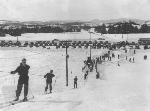A vintage black-and-white photo of skiers on a snowy slope with cars parked in the background and mountains in the distance.