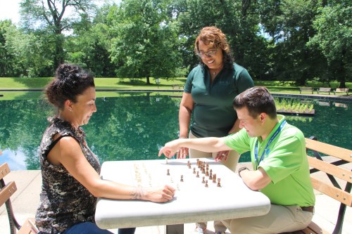 Two people play chess outdoors while a third person observes, surrounded by greenery and a pond.