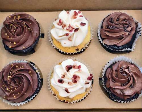 A box of six decorative cupcakes with chocolate and vanilla frosting, some topped with rose petals and gold sprinkles.