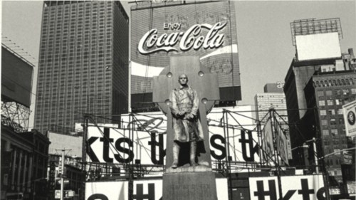 Black and white image of a statue in a city square, with a large Coca-Cola advertisement in the background.