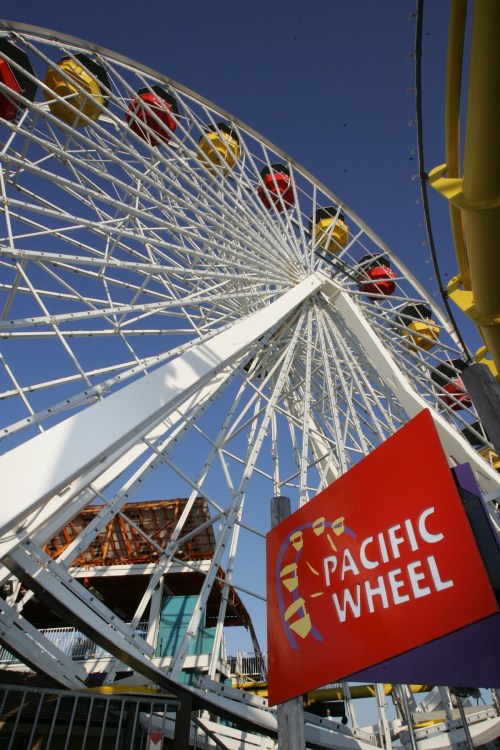 A close-up of the Pacific Wheel ferris wheel against a clear blue sky, with colorful gondolas and a red sign.