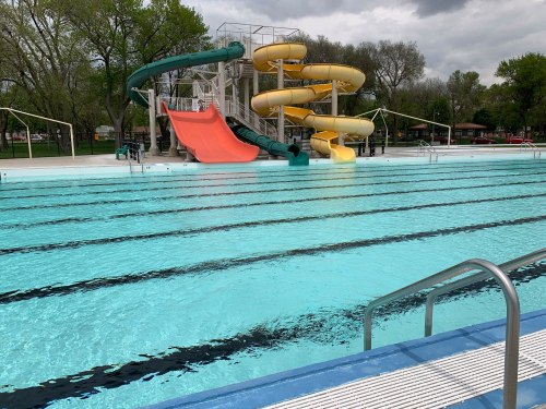 A swimming pool with colorful water slides and clear blue water, surrounded by trees under a cloudy sky.