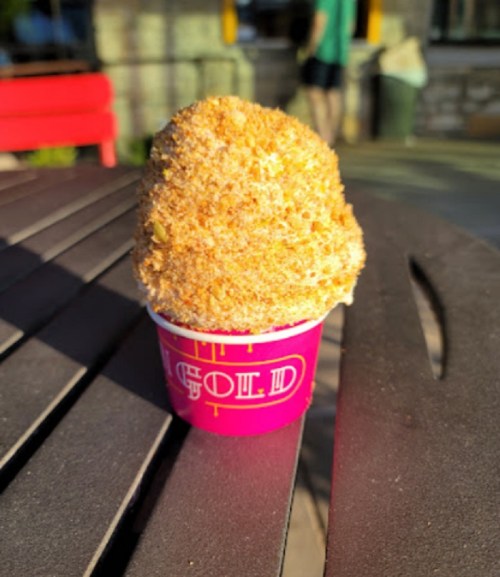A small cup of dessert topped with a golden, crumbly coating, sitting on a wooden table in a sunny outdoor setting.