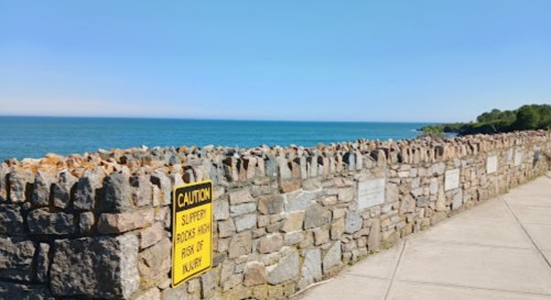 A stone wall along a coastal path with a caution sign, overlooking a calm blue sea under a clear sky.