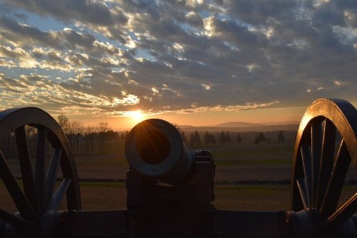 A cannon silhouette against a sunset, with clouds and distant hills in the background.