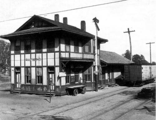 Historic train station with a two-story building, wooden structure, and freight cars nearby on a dirt road.