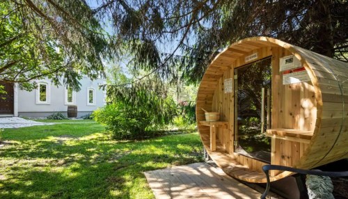 A wooden sauna in a lush garden, surrounded by trees and greenery, with a sunny blue sky above.