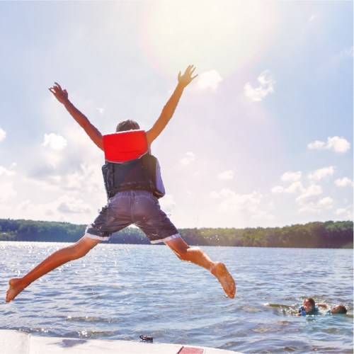 A child jumps off a boat into a lake on a sunny day, with friends swimming nearby.