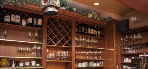 A wooden wine rack filled with various bottles, surrounded by glasses and decorative greenery on the shelves.