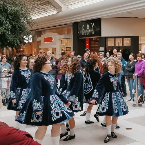 A group of dancers in blue dresses perform in a mall, with an audience watching in the background.