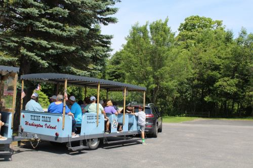 A tour train with passengers waits near a tree-lined area, with a car parked beside it on a sunny day.